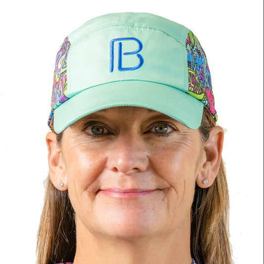 Woman smiles wearing a mint green five-panel running hat with a colorful doodle pattern and a blue "B" logo.
