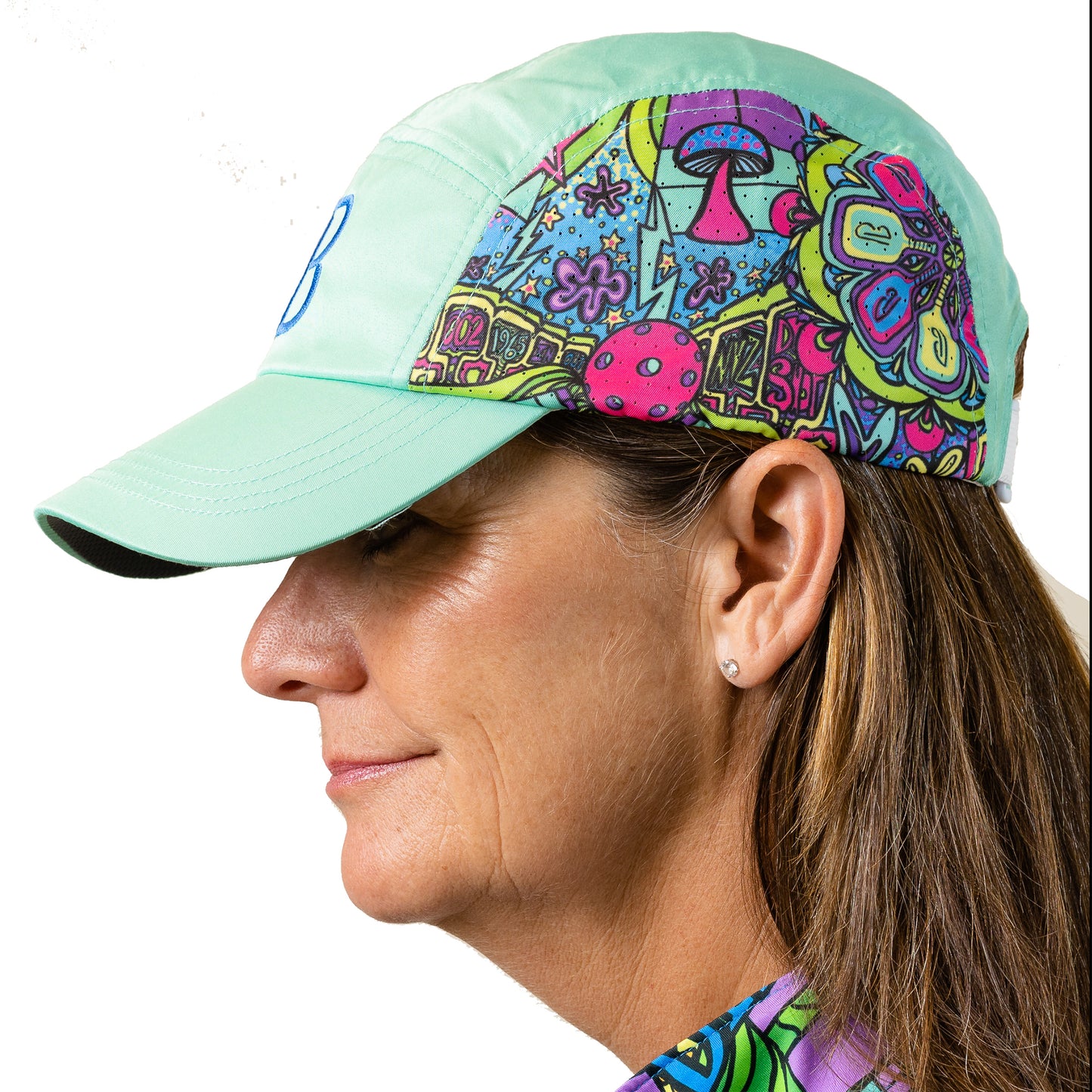 Woman models a mint green performance running hat with a colorful psychedelic mushroom print and breathable mesh panels.
