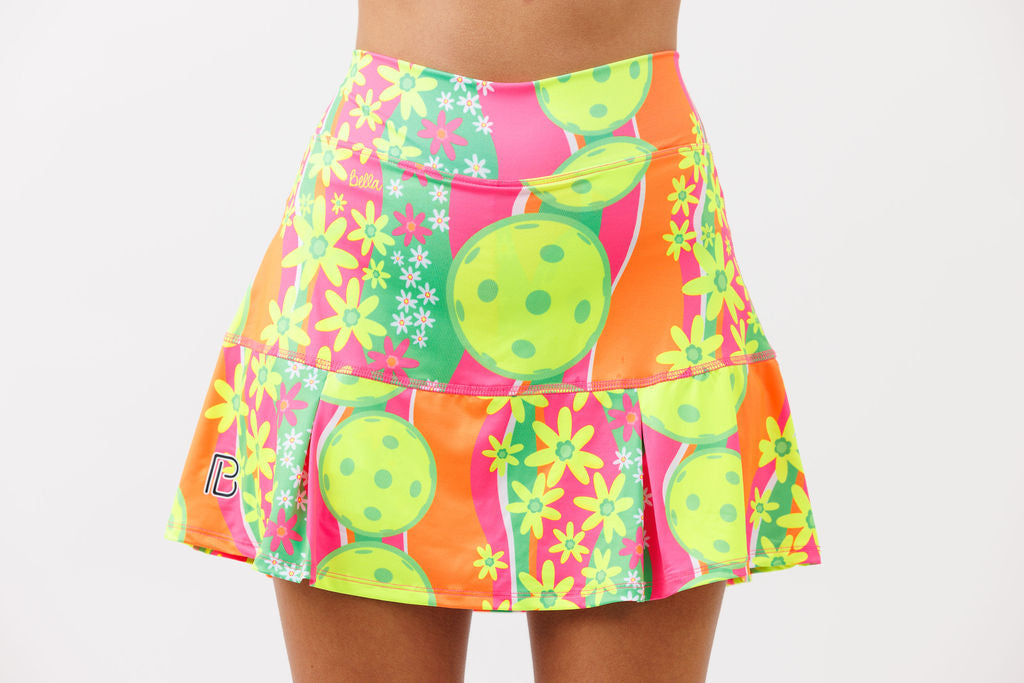 Woman models a pleated athletic skirt featuring a pink, orange, and green floral print with pickleball graphics.