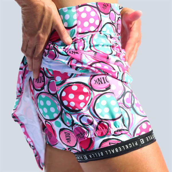 Woman models white athletic skirt with pink and green pickleball print and built-in shorts.