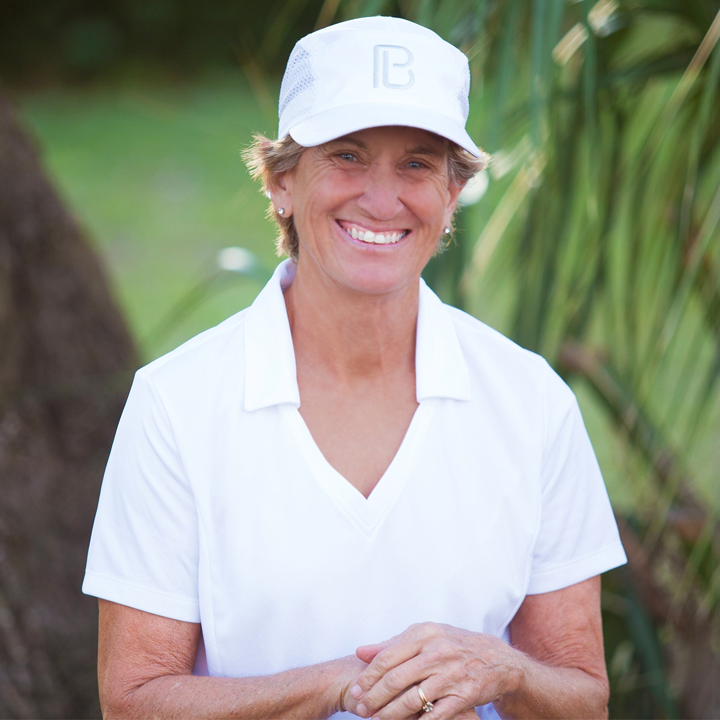 Woman smiles brightly in a white polo and white baseball cap while standing outdoors.