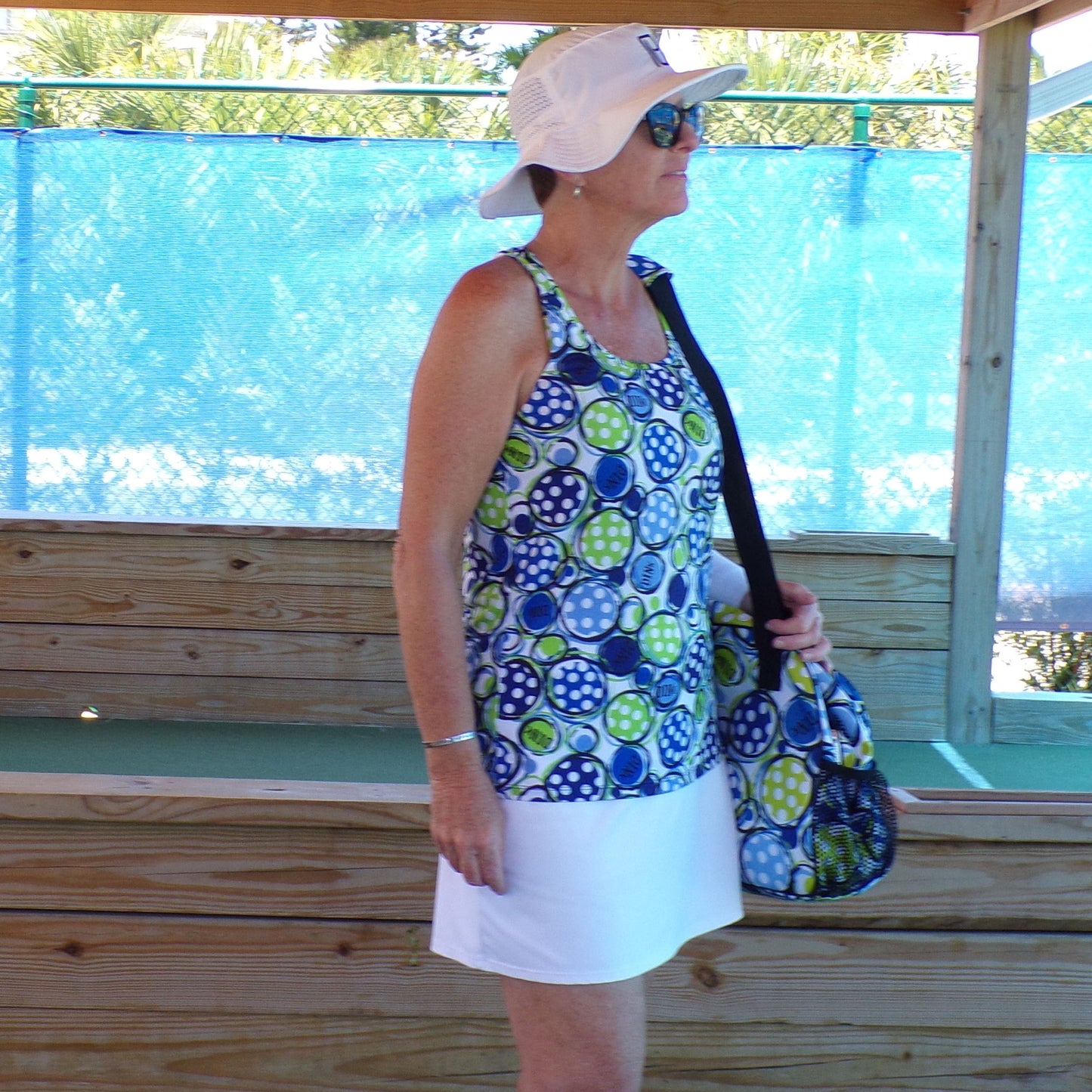 Woman models a white tennis skort and a white, blue, and green polka dot athletic top. She holds a matching polka dot bag over her shoulder.