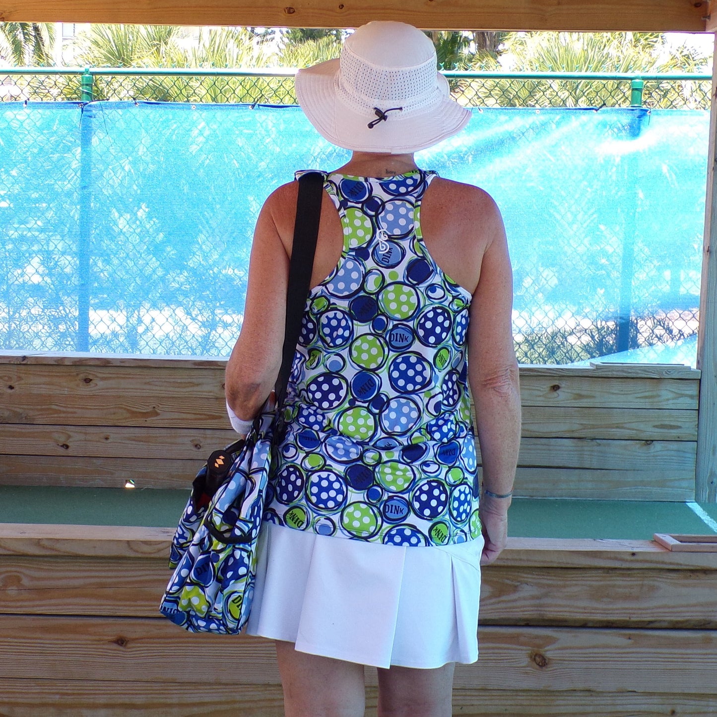 Woman wearing a blue and green polka dot athletic top and white skort watches a pickleball match from behind a wooden fence.