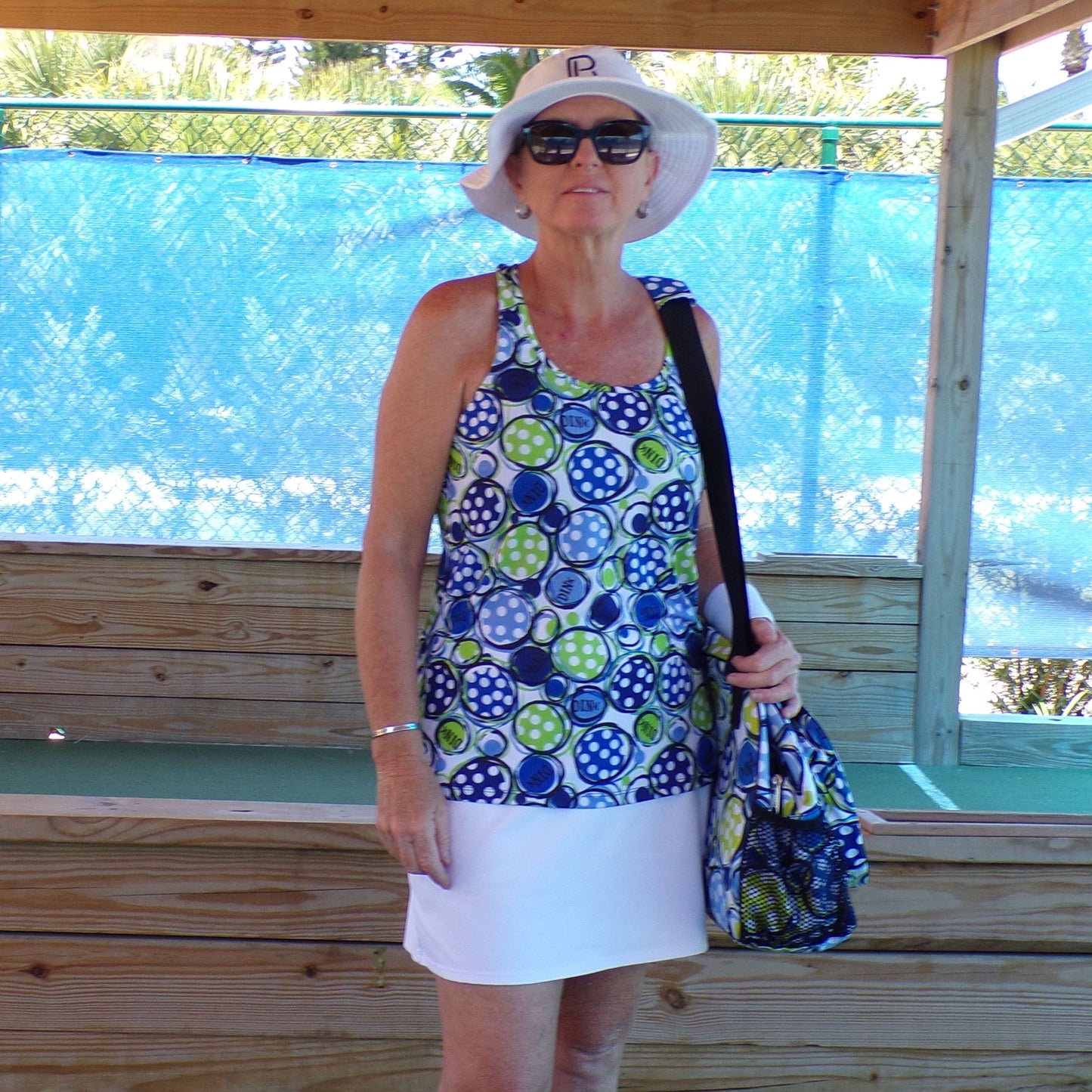 Woman wears a white and blue polka dot tennis dress and a white sun visor while standing on a wooden pickleball court.