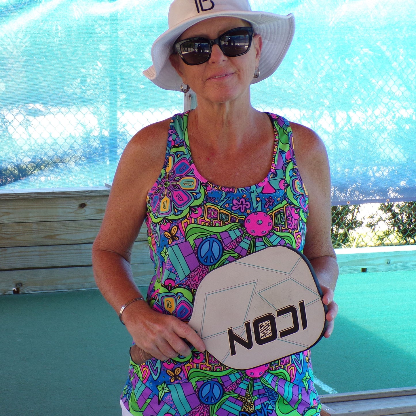 Woman in colorful athletic wear holds a white and black ICON pickleball paddle in front of a blue fence.