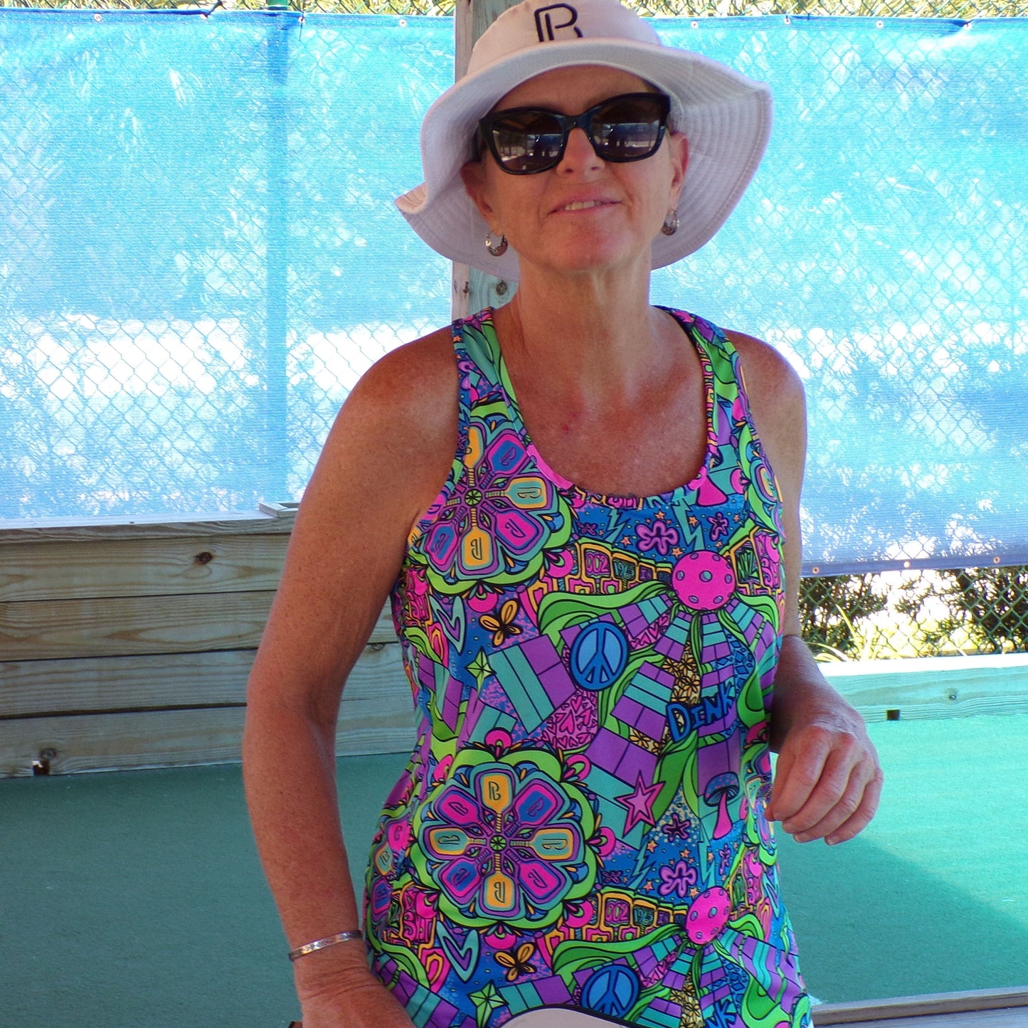 Woman wearing a brightly colored psychedelic print tank top and white sun hat smiles outdoors.