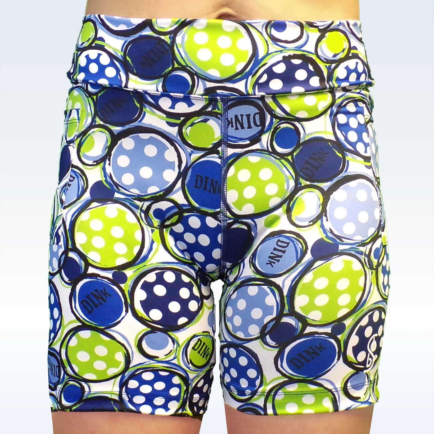 Woman models athletic shorts featuring a white, blue, and green pattern of pickleballs with the word "DINK."