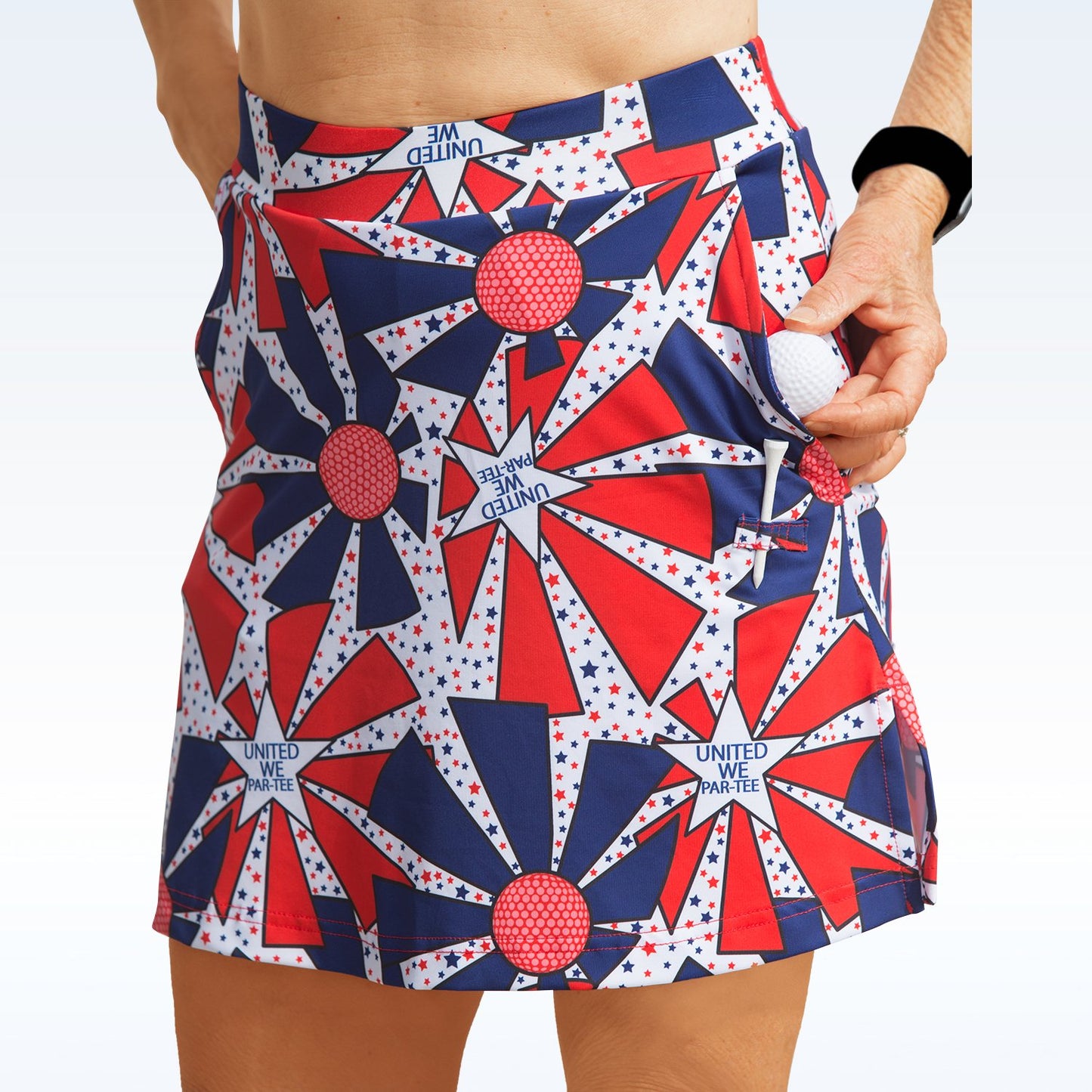 Woman models a red, white, and blue golf skort with a starburst pattern and places a golf ball in the side pocket.