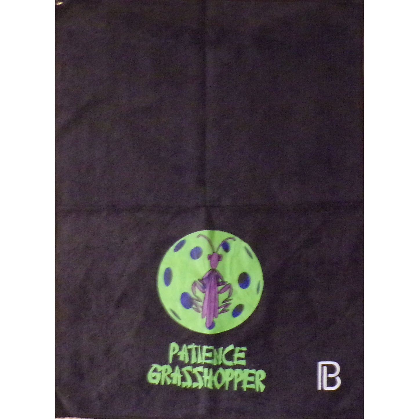 A black pickleball towel displays a neon green pickleball with a purple praying mantis at its center. Below the design, the words "PATIENCE GRASSHOPPER" are printed in bold, green letters.