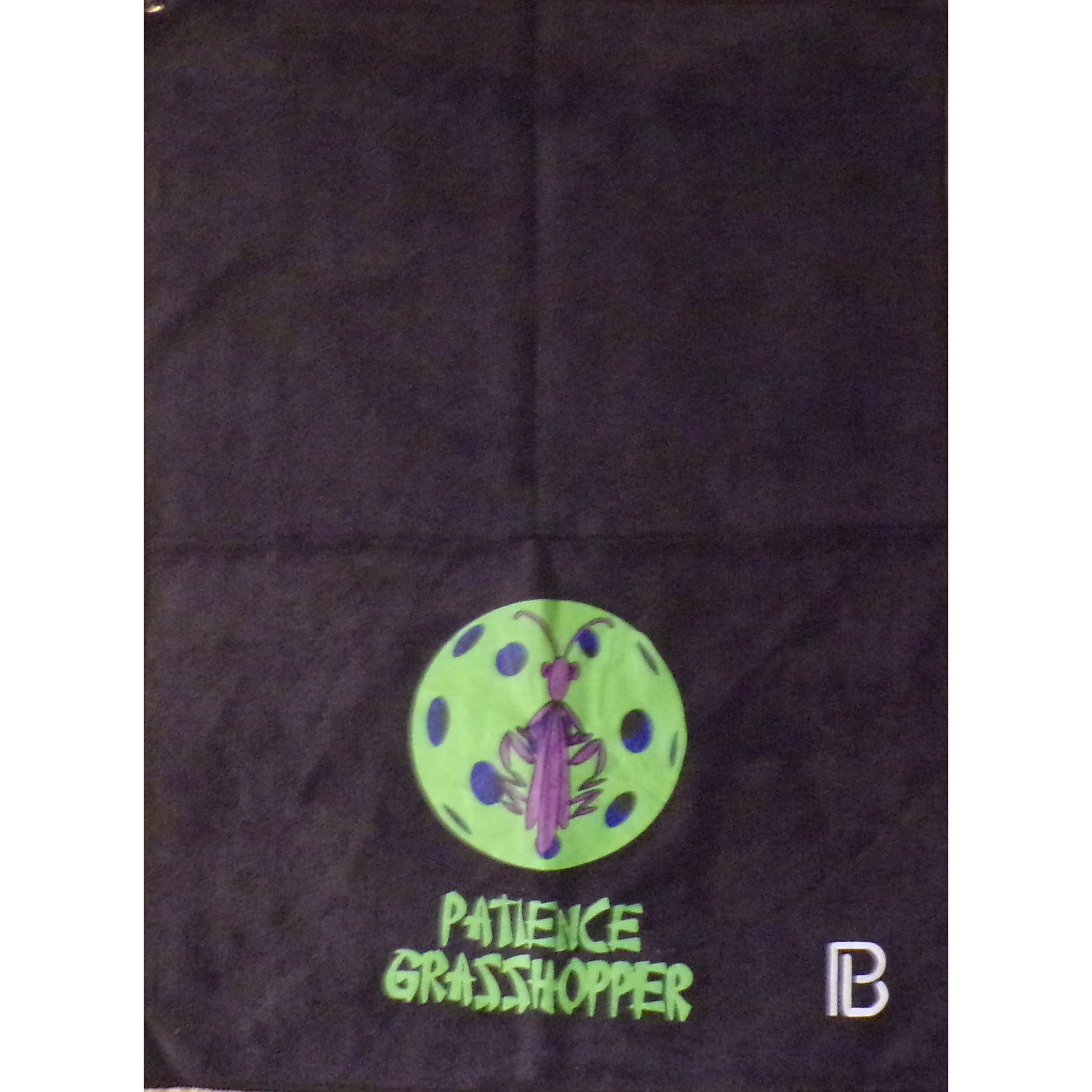 A black pickleball towel displays a neon green pickleball with a purple praying mantis at its center. Below the design, the words "PATIENCE GRASSHOPPER" are printed in bold, green letters.