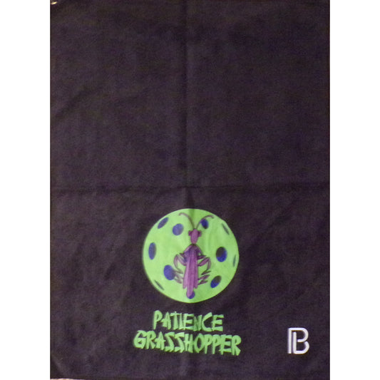 A black pickleball towel displays a neon green pickleball with a purple praying mantis at its center. Below the design, the words "PATIENCE GRASSHOPPER" are printed in bold, green letters.