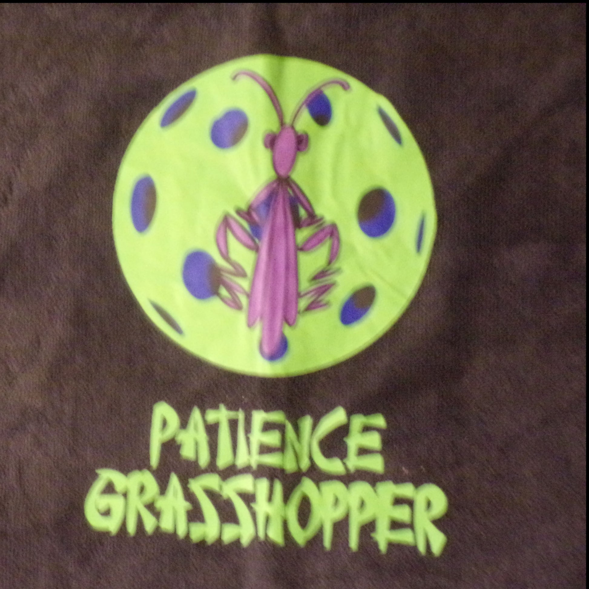 A purple praying mantis stands on a lime-green pickleball. The text "PATIENCE GRASSHOPPER" is printed below the image.