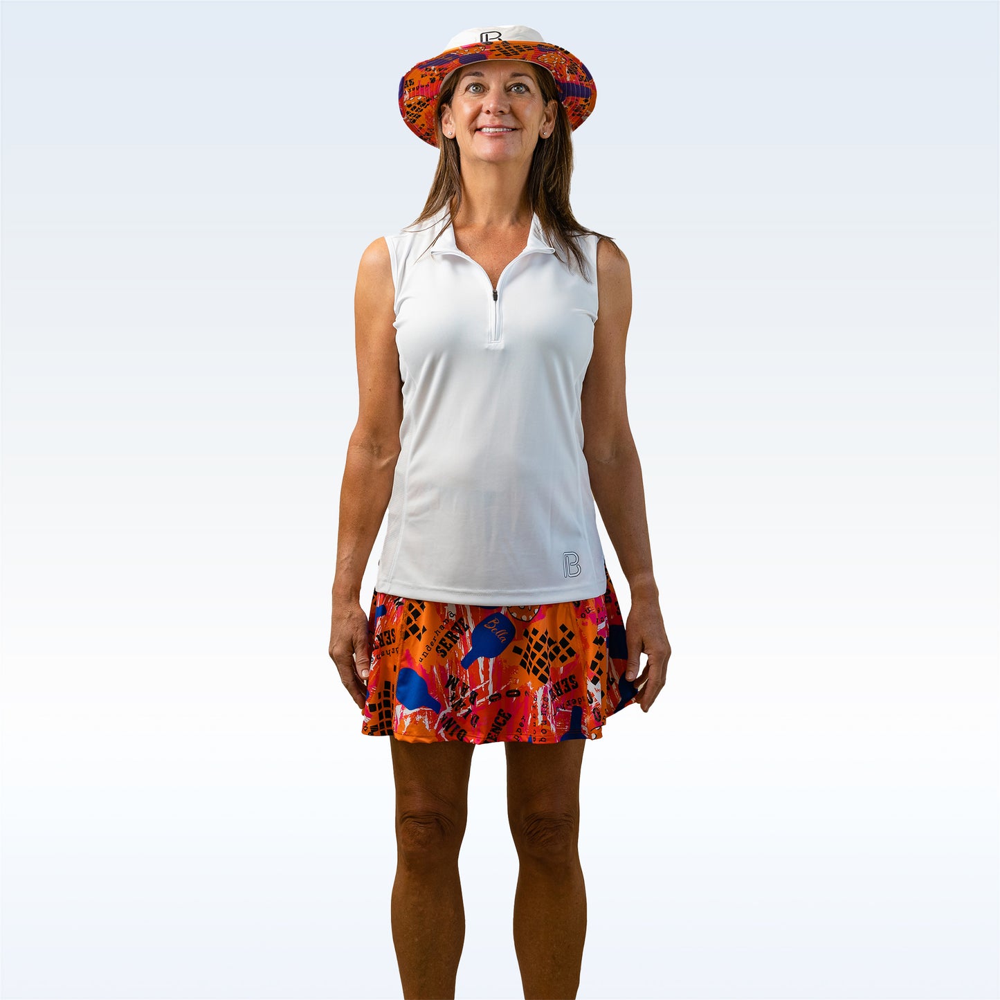 Woman models white golf shirt and orange patterned golf skort with white bucket hat. 
